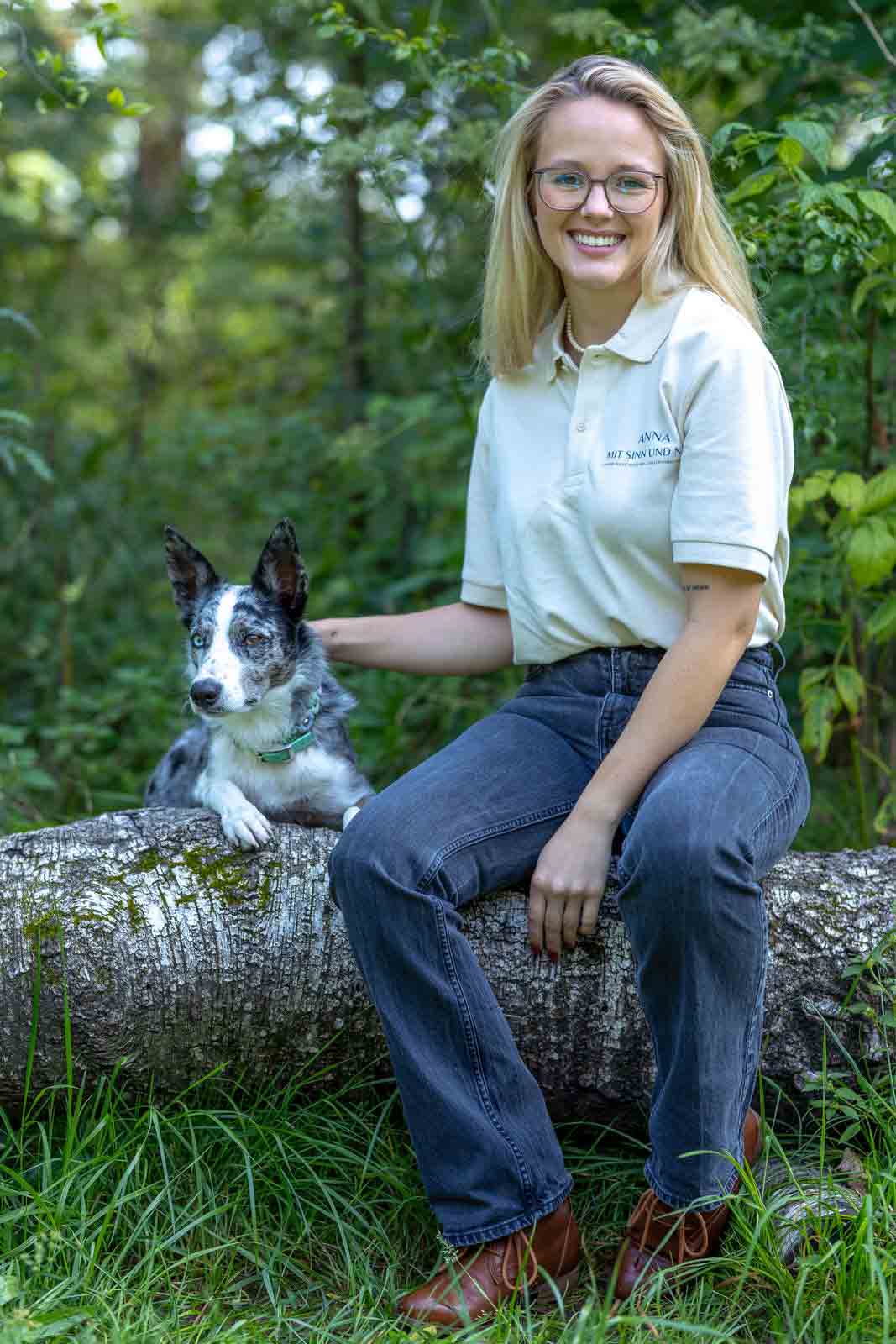 Anna von Mit Sinn und Napf sitzt auf einem Baumstamm, streichelt ihren Hund und schaut den Betrachter an.